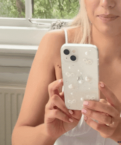 A person with long blonde hair is holding a Coconut Lane Coquette Dreams Phone Case in their right hand. They sit near a window with green foliage visible outside, wearing a white sleeveless top and a ring on their left hand.