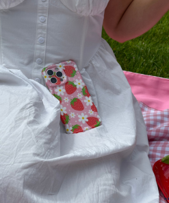 A person wearing a white dress sits on a pink and white checkered blanket on the grass. An extra protective smartphone with the Coconut Lane's Wavy Phone Case - Berry Cute Strawberry rests on their dress. Nearby, a red heart-shaped purse and a beaded accessory are visible.
