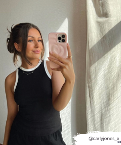 A woman taking a mirror selfie with her smartphone, adorned in the Coconut Lane Melting Heart Phone Case - Peony Pink. She is smiling with her hair styled in a bun with loose strands and is wearing a sleeveless black top with white trim. Her Instagram handle, @carlyjones_x, appears in the bottom right corner.