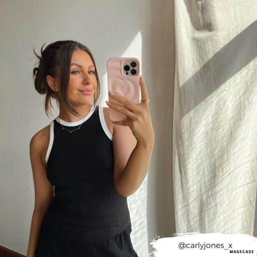 A woman taking a mirror selfie with her smartphone, adorned in the Coconut Lane Melting Heart Phone Case - Peony Pink. She is smiling with her hair styled in a bun with loose strands and is wearing a sleeveless black top with white trim. Her Instagram handle, @carlyjones_x, appears in the bottom right corner.