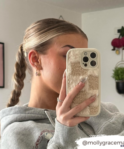A person with a braided hairstyle takes a mirror selfie, showcasing the back of their phone adorned with the Cosy Teddy Phone Case - Vanilla Biscuit by Coconut Lane in beige and white checks. They are wearing a gray hoodie in a room decorated with various items, including a painting and hanging decor. The text 
