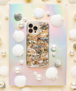 A smartphone with a colorful, iridescent case featuring an abstract design sits on a holographic surface. The phone, adorned with a Real Natural Shell Phone Case - Black from Coconut Lane, is surrounded by white seashells, pearls, and small textured stones, creating an ocean-inspired aesthetic.