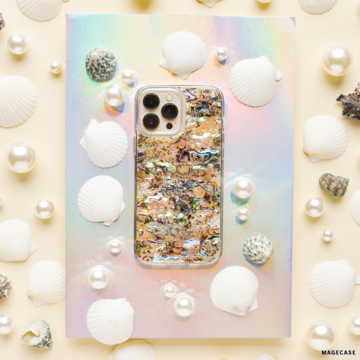 A smartphone with a colorful, iridescent case featuring an abstract design sits on a holographic surface. The phone, adorned with a Real Natural Shell Phone Case - Black from Coconut Lane, is surrounded by white seashells, pearls, and small textured stones, creating an ocean-inspired aesthetic.