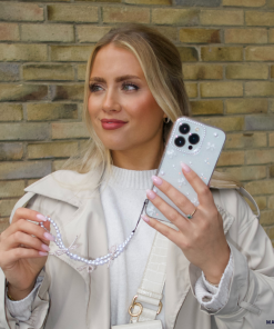 A woman with long blonde hair is holding a smartphone featuring the Mini White Bows Phone Case from Coconut Lane, along with a beaded strap. She is dressed in a light-colored coat and sweater, set against a brick wall backdrop. Her nails are painted light pink, and she seems to be gazing slightly off-camera.