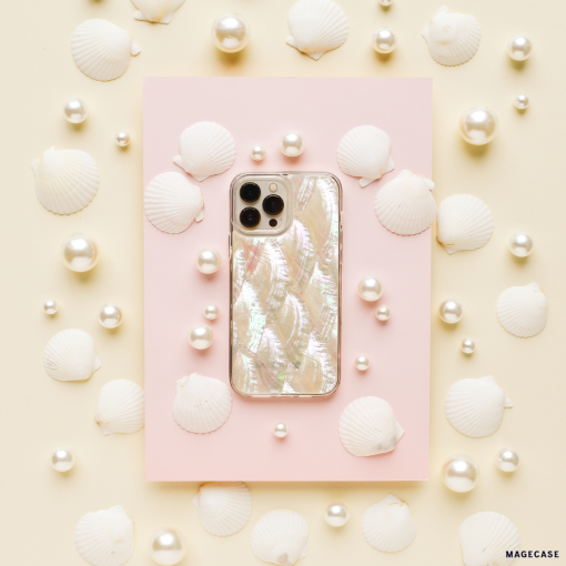 A white iPhone adorned with Coconut Lane's Real Natural Shell Phone Case is set on a pastel pink rectangular card, surrounded by white seashells and pearls, all against a cream-colored background. This arrangement creates a marine-themed aesthetic with a soft, elegant feel.