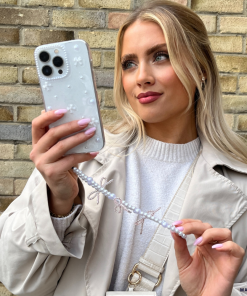 A woman with long blonde hair holds up her smartphone, encased in a Coconut Lane Mini White Bows Phone Case, to take a selfie. She is wearing a light tan coat over a white top. Her phone’s beaded strap complements the clear design of the iPhone 15 case. The background features a brick wall.