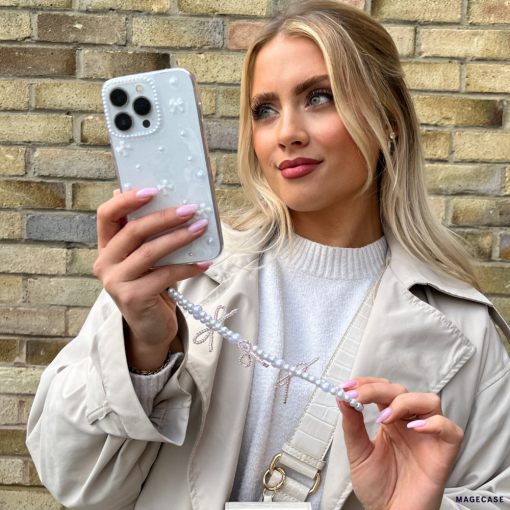 A woman with long blonde hair holds up her smartphone, encased in a Coconut Lane Mini White Bows Phone Case, to take a selfie. She is wearing a light tan coat over a white top. Her phone’s beaded strap complements the clear design of the iPhone 15 case. The background features a brick wall.