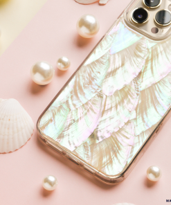 A smartphone with a shimmering, iridescent case designed to resemble fish scales lies on a pink and yellow background. The scene, featuring the Real Natural Shell Phone Case in White by Coconut Lane, is decorated with pearls and small white seashells, creating a delicate, ocean-themed aesthetic.