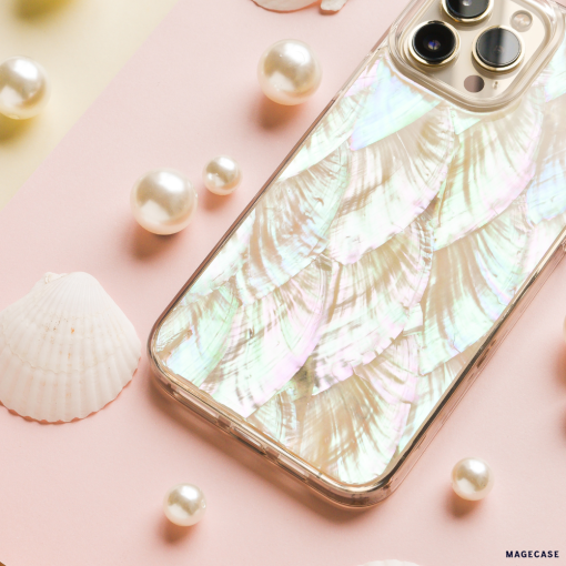 A smartphone with a shimmering, iridescent case designed to resemble fish scales lies on a pink and yellow background. The scene, featuring the Real Natural Shell Phone Case in White by Coconut Lane, is decorated with pearls and small white seashells, creating a delicate, ocean-themed aesthetic.