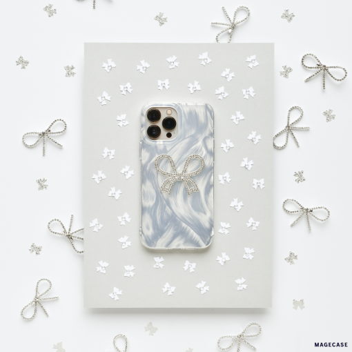 A Coconut Lane Diamante Bow Phone Case, featuring a marbled white and gray design with an extra protective decorative bow, is placed on a gray sheet surrounded by small paper stars and silver wire bows. The white background adds to the clean and elegant aesthetic.