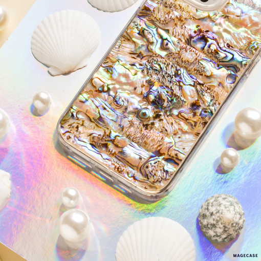 A smartphone with a reflective Coconut Lane Real Natural Shell Phone Case in Black lies on a pastel-colored, iridescent surface. Surrounding the phone are authentic durable shells and variously sized pearls, enhancing the aquatic theme.