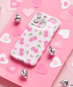 A Wavy Phone Case - Disco Cherries from Coconut Lane lies on a pink surface adorned with various paper hearts and small silvery disco balls. The case, featuring a playful cherry pattern on a white background, exudes a whimsical, romantic vibe.