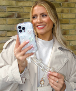 A blonde woman with a bright smile holds an iPhone 15 adorned with Coconut Lane's clear Pearly Hearts Phone Case and a pearl chain. She is leaning against a brick wall, wearing a light beige trench coat over a white sweater, accessorized with a small white handbag and a ring on her finger.