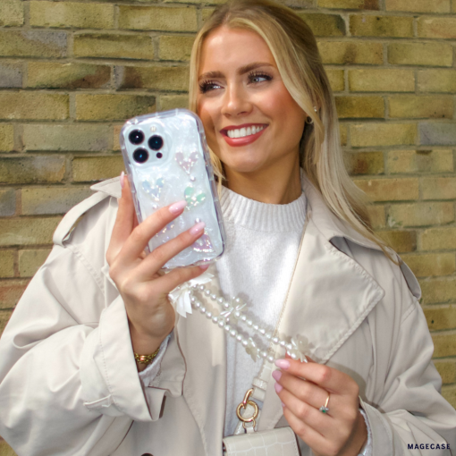 A blonde woman with a bright smile holds an iPhone 15 adorned with Coconut Lane's clear Pearly Hearts Phone Case and a pearl chain. She is leaning against a brick wall, wearing a light beige trench coat over a white sweater, accessorized with a small white handbag and a ring on her finger.