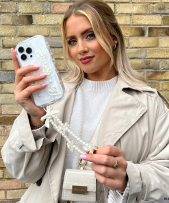 A woman with long blonde hair is holding a smartphone adorned with Coconut Lane's Pearly Hearts Phone Case - Clear, featuring a beaded strap. She is wearing a beige trench coat over a white sweater and has a small white handbag hanging from her shoulder. Standing in front of a brick wall, she poses confidently for the camera.