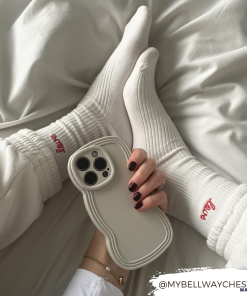 A person with dark nail polish holds a lightweight, vanilla-colored phone. Their legs are crossed, showcasing white socks with 