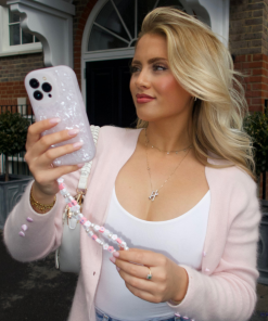 A woman with blonde hair, dressed in a pink cardigan and white top, holds up her smartphone encased in a Coconut Lane Moonstone Pink Phone Case to take a selfie. She stands in front of a brick building with arched windows and decorative plants, smiling slightly as she looks at the phone's screen.