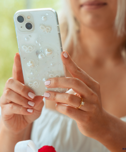 A person with light pink manicured nails is holding an iPhone 15 with a Coquette Dreams Phone Case by Coconut Lane, adorned with white pearls. They are wearing a white top, and a delicate gold ring with a clear gemstone is visible on their left hand. A blurred green background provides a serene backdrop.