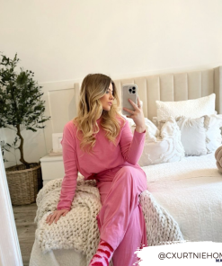 A woman with long, wavy blonde hair sits on a bed in a light, cozy bedroom. She is wearing a pink lounge set and holding a Curvy Phone Case - Cream from Coconut Lane in her left hand. The bed is decorated with white pillows and a chunky knit blanket. A potted plant is in the background. Text on the image reads 