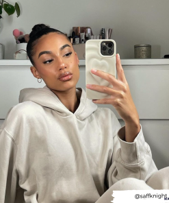 A woman with a neatly tied bun takes a mirror selfie while wearing a beige hoodie. She has a calm expression and is holding her phone, protected by the stylish Melting Heart Phone Case in Coconut Cream from Coconut Lane. The background includes a plant, books, and neatly arranged items on a white shelf. Username 