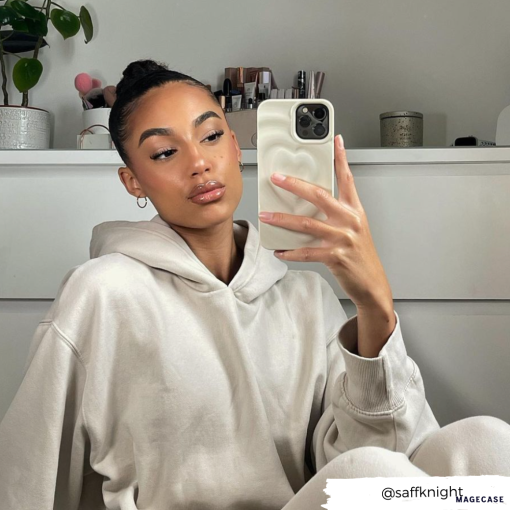 A woman with a neatly tied bun takes a mirror selfie while wearing a beige hoodie. She has a calm expression and is holding her phone, protected by the stylish Melting Heart Phone Case in Coconut Cream from Coconut Lane. The background includes a plant, books, and neatly arranged items on a white shelf. Username "@saffknight" is visible.