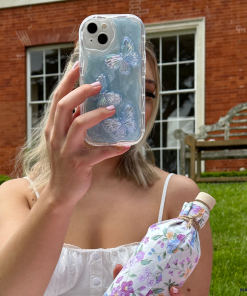 A person with blonde hair, wearing a white tank top, holds up a Shimmer Blue Butterflies Phone Case by Coconut Lane, partially obscuring their face. In their other hand, they hold a floral-patterned water bottle. They are in front of a brick building and sitting on grass.