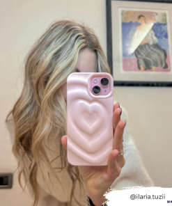 A person with long, wavy blonde hair takes a mirror selfie while holding a phone encased in the Coconut Lane Melting Heart Phone Case in Peony Pink, which features a 3D melting heart design. A piece of art is visible on the wall behind them. The Instagram handle @ilaria.tuzii is written in the bottom right corner.