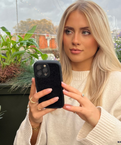 A woman with long blonde hair and a light-colored sweater is sitting outdoors, holding a smartphone with the Cosy Teddy Phone Case - Black from Coconut Lane. She appears focused, with greenery and potted plants in the background. She's wearing several rings and a bracelet, with a calm expression on her face.