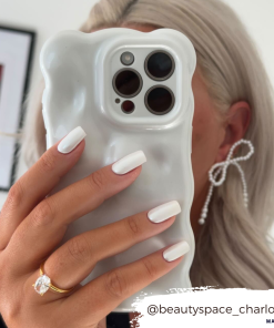 A person with platinum blonde hair is holding a Coconut Lane 3D Pearl Phone Case - White, featuring a wavy design and pearl white effect, obscuring most of their face. They have white manicured nails, a diamond ring on their ring finger, and are wearing bow-shaped earrings. The Instagram handle @beautyspace_charlotte is visible.