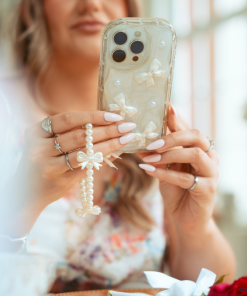 A woman with manicured nails holds the Pearls & Bows Phone Case by Coconut Lane, adorned with pearls and bows. She wears rings and a bracelet that match the phone's decoration. The background is softly blurred, focusing on her hands and the protective phone case.