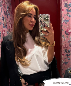 A person with long, wavy brown hair takes a mirror selfie using a Coconut Lane 3D Chrome Phone Case. They are wearing a white shirt and a watch, standing in front of a red door and floral wallpaper. Their Instagram handle "@XMEVELYNT" is overlaid at the bottom right corner.