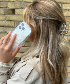 A woman with long, wavy blonde hair holds her iPhone15 adorned with the Pearly Hearts Phone Case - Clear by Coconut Lane to her ear. She is wearing a beige trench coat and a pearl bracelet. Her hair is accessorized with a decorative hair clip. The background features a brick wall, adding to the chic ambiance.