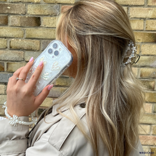 A woman with long, wavy blonde hair holds her iPhone15 adorned with the Pearly Hearts Phone Case - Clear by Coconut Lane to her ear. She is wearing a beige trench coat and a pearl bracelet. Her hair is accessorized with a decorative hair clip. The background features a brick wall, adding to the chic ambiance.