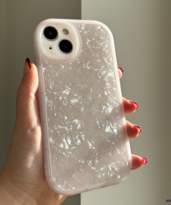 A hand with red-painted nails holds a Coconut Lane Moonstone Pink Phone Case for the iPhone15. The iridescent, protective case features a textured and translucent design that catches the light, creating a shimmering effect. The modestly lit background highlights the intricate details of the phone case.