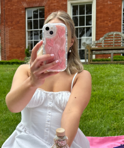 A woman in a white dress sits on a lawn in front of a brick building, holding her phone up to take a selfie. The Coconut Lane Shimmer Pink Hearts Phone Case adds an extra layer of protection while displaying glittery pink hearts. A pink picnic blanket and a small bottle are visible in the foreground.