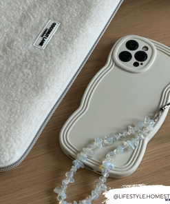 A white, textured laptop sleeve and a lightweight smartphone with a uniquely shaped Coconut Lane NAKD Curvy Phone Case - Vanilla lie on a wooden surface. The phone case features a beaded strap attached. The text 