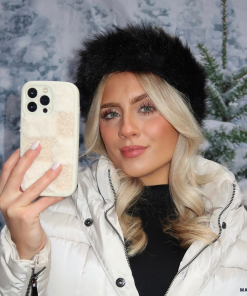 A person with long blonde hair takes a selfie with their smartphone, which is encased in the Cosy Teddy Phone Case - Vanilla Biscuit from Coconut Lane. They are wearing a white quilted jacket and a black fur hat, standing against a snowy winter background with trees.
