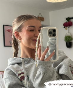 A person in a grey hoodie takes a mirror selfie using a phone protected by the stylish Curvy Mirror Phone Case - Cream from Coconut Lane. They are winking and smiling, with their hair styled in a braid. The room behind them includes a bed, wall art, and hanging decorations. The username 