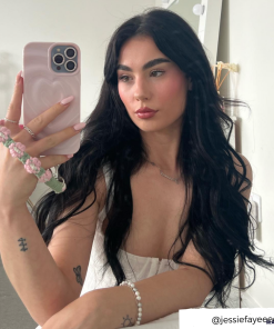 A person with long, wavy dark hair takes a mirror selfie using a phone with Coconut Lane's Melting Heart Phone Case in Peony Pink. They are wearing a white top and have tattoos on their left arm. A bracelet and phone charm featuring pink and white beads are visible. The Instagram handle 
