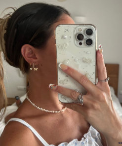 A woman with a ponytail takes a mirror selfie, partially covering her face with her Coconut Lane Coquette Dreams Phone Case. She sports butterfly-shaped earrings, a pearl necklace, and flaunts a ring-adorned hand with manicured nails painted in a floral design. Her iPhone15 case stands out, adding an extra protective charm to the bedroom snapshot.