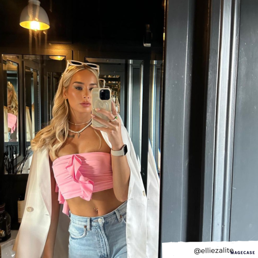 A woman with long blonde hair is taking a mirror selfie. She is wearing a pink strapless top with a bow, light denim jeans, and a white blazer draped over her shoulders. Sunglasses rest on her head as she holds her phone in its Melting Heart Phone Case - Coconut Cream by Coconut Lane. The Instagram handle @elliezalite is displayed in the bottom right corner.