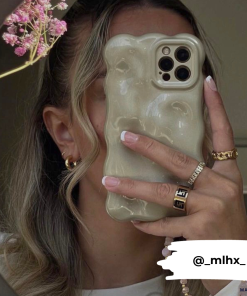 A person with wavy hair holds an iPhone 15 featuring the Coconut Lane 3D Pearl Phone Case - Gold in front of their face. They wear gold hoop earrings, a gold ring, and have manicured nails. There are pink flowers visible in the upper left corner. The Instagram handle @_mlhx_ is displayed at the bottom.