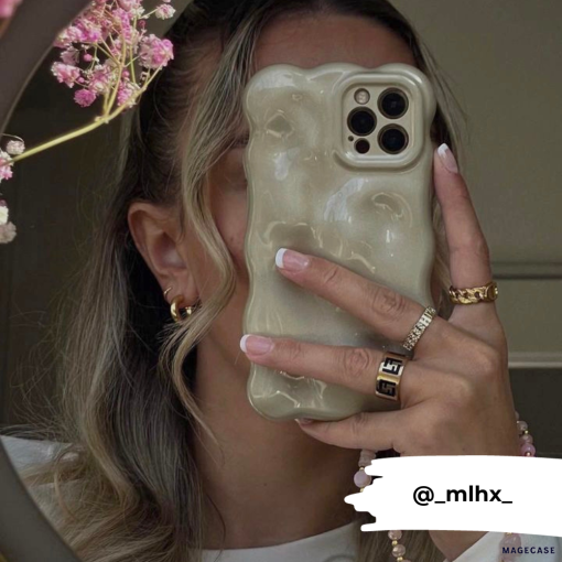 A person with wavy hair holds an iPhone 15 featuring the Coconut Lane 3D Pearl Phone Case - Gold in front of their face. They wear gold hoop earrings, a gold ring, and have manicured nails. There are pink flowers visible in the upper left corner. The Instagram handle @_mlhx_ is displayed at the bottom.