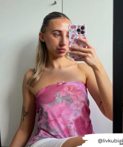 A person with long blonde hair takes a mirror selfie. They are wearing a pink strapless top with a floral pattern and hold a phone featuring Coconut Lane's extra protective Wavy Phone Case - Disco Cherries. A tattoo is visible on their right forearm, and the username 