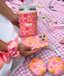 A person is holding the Coconut Lane Wavy Phone Case - Pink & Orange Bloom, seated on a pink checkered picnic blanket. The scene features matching floral accessories like a scrunchie, drink holder, headphones, and various items including a pearl necklace and eyeglasses, all in a coordinating pink theme.