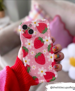 A hand with colorful, strawberry-themed nail art holds a phone encased in the extra-protective Coconut Lane Wavy Phone Case - Berry Cute Strawberry. The background showcases a blurred view of a cozy room with soft furnishings, including a pillow and a small table with a plant.
