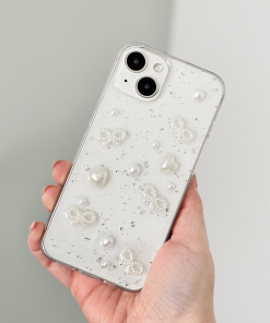 A hand holds an iPhone15 with a Coconut Lane Coquette Dreams Phone Case. The decorative case features embedded pearls, butterfly shapes, and sparkling embellishments on a translucent background, offering extra protective qualities. The phone has dual cameras and a sleek, glossy finish. The background is blurred.