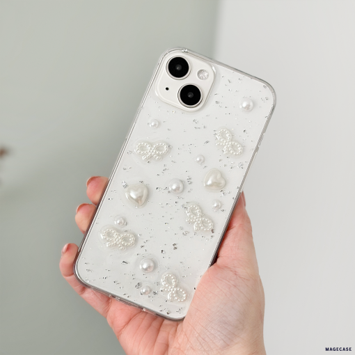 A hand holds an iPhone15 with a Coconut Lane Coquette Dreams Phone Case. The decorative case features embedded pearls, butterfly shapes, and sparkling embellishments on a translucent background, offering extra protective qualities. The phone has dual cameras and a sleek, glossy finish. The background is blurred.