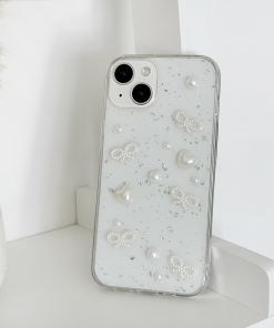 A silver smartphone adorned with the Coquette Dreams Phone Case from Coconut Lane, featuring elegant pearls and bead designs, stands upright on a white shelf. In the background, a vase filled with beige dried grasses adds to the serene setting. The iPhone15 Case boasts two rear cameras, enhancing the minimalistic and stylish arrangement.