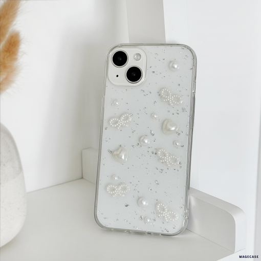 A silver smartphone adorned with the Coquette Dreams Phone Case from Coconut Lane, featuring elegant pearls and bead designs, stands upright on a white shelf. In the background, a vase filled with beige dried grasses adds to the serene setting. The iPhone15 Case boasts two rear cameras, enhancing the minimalistic and stylish arrangement.